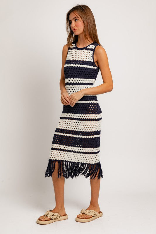 A woman in the Sleeveless Crochet Effect Sweater Dress, featuring black and white stripes with a fringe hem, holds a textured beige handbag. She pairs the outfit with beige sandals, standing against a plain white background and exuding an effortlessly chic vibe.