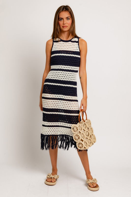 A woman in the Sleeveless Crochet Effect Sweater Dress, featuring black and white stripes with a fringe hem, holds a textured beige handbag. She pairs the outfit with beige sandals, standing against a plain white background and exuding an effortlessly chic vibe.