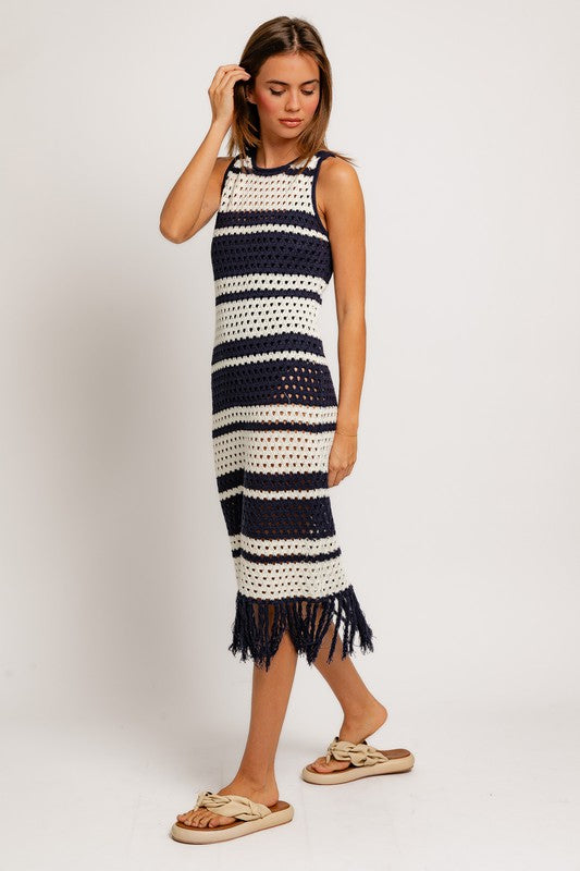 A woman in the Sleeveless Crochet Effect Sweater Dress, featuring black and white stripes with a fringe hem, holds a textured beige handbag. She pairs the outfit with beige sandals, standing against a plain white background and exuding an effortlessly chic vibe.