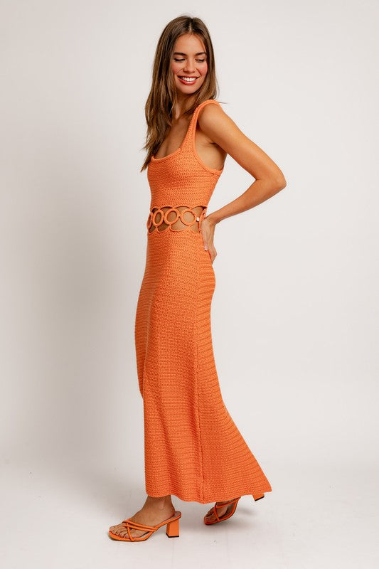 A person stands against a plain background wearing a Square Neck Sleeveless Crochet Midi Dress in beige, featuring circular cutouts at the waist. They have long hair and are wearing sandals.