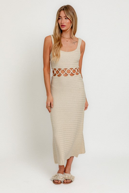 A person stands against a plain background wearing a Square Neck Sleeveless Crochet Midi Dress in beige, featuring circular cutouts at the waist. They have long hair and are wearing sandals.