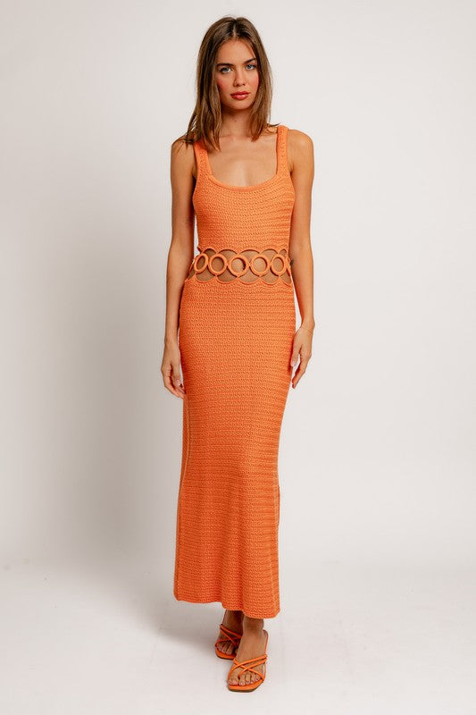 A person stands against a plain background wearing a Square Neck Sleeveless Crochet Midi Dress in beige, featuring circular cutouts at the waist. They have long hair and are wearing sandals.