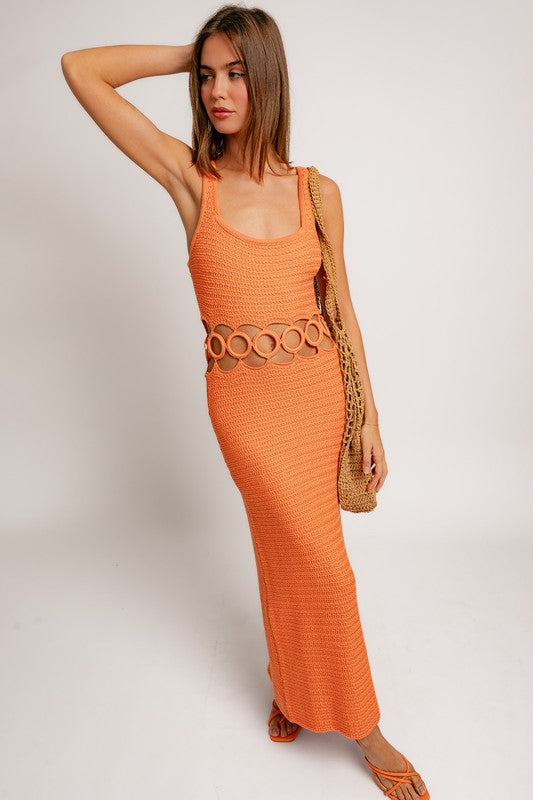 A person stands against a plain background wearing a Square Neck Sleeveless Crochet Midi Dress in beige, featuring circular cutouts at the waist. They have long hair and are wearing sandals.