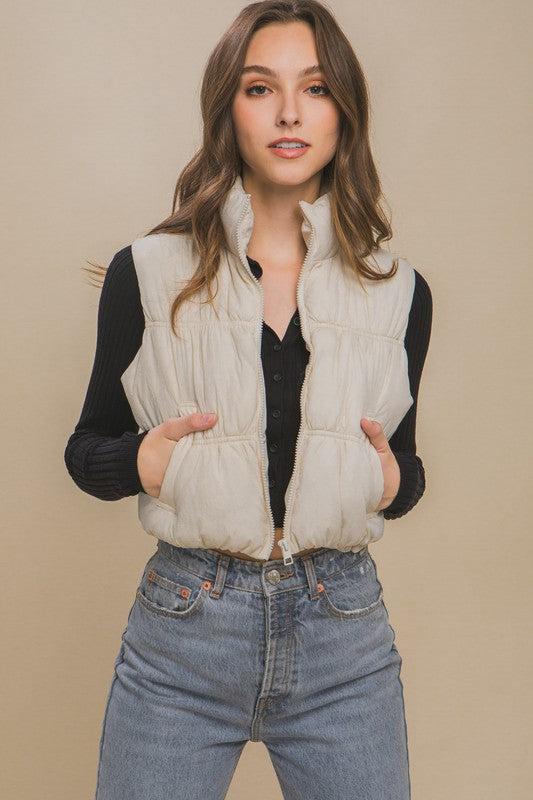 A woman with long hair is smiling, dressed in a light gray High Neck Puffer Vest, a black long-sleeve top, and high-waisted blue jeans. She is standing against a beige background showcasing her winter style.