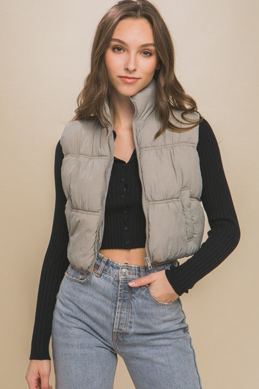 A woman with long hair is smiling, dressed in a light gray High Neck Puffer Vest, a black long-sleeve top, and high-waisted blue jeans. She is standing against a beige background showcasing her winter style.