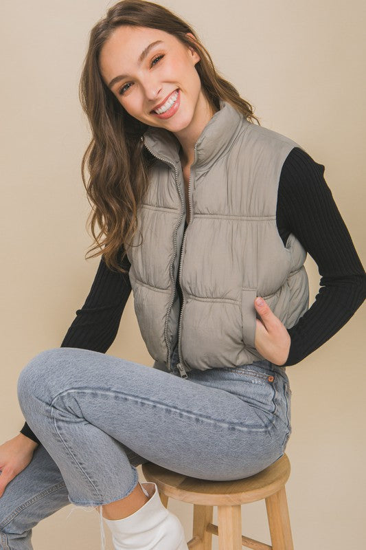 A woman with long hair is smiling, dressed in a light gray High Neck Puffer Vest, a black long-sleeve top, and high-waisted blue jeans. She is standing against a beige background showcasing her winter style.