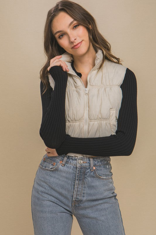 A woman with long hair is smiling, dressed in a light gray High Neck Puffer Vest, a black long-sleeve top, and high-waisted blue jeans. She is standing against a beige background showcasing her winter style.