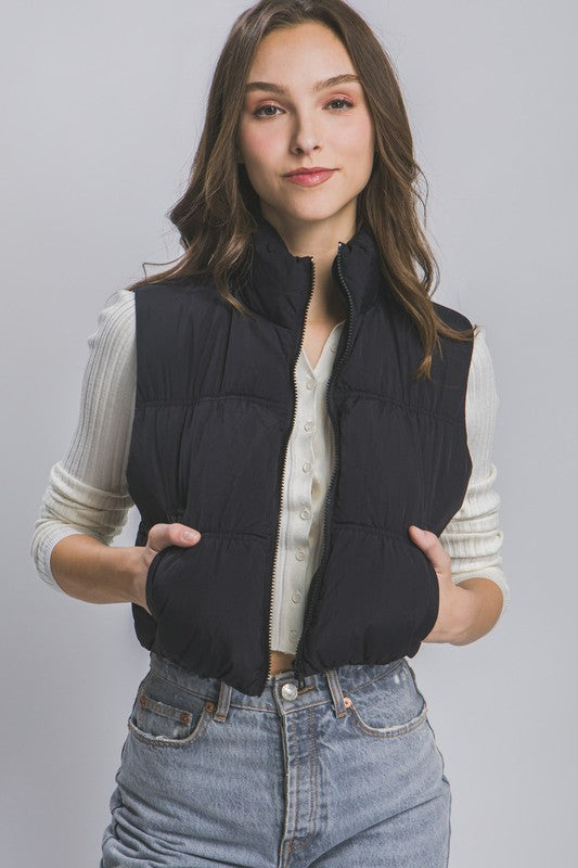 A woman with long hair is smiling, dressed in a light gray High Neck Puffer Vest, a black long-sleeve top, and high-waisted blue jeans. She is standing against a beige background showcasing her winter style.