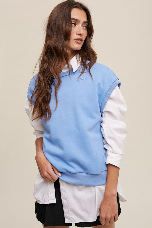 A woman stands against a neutral background, wearing the Short Sleeve Boxy Crew Neck Sweat Top in blue with ribbed detailing over a white button-up shirt.