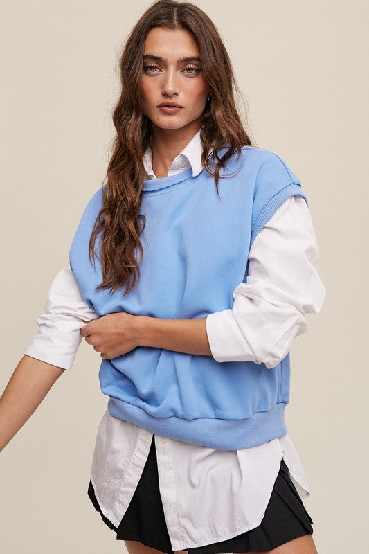 A woman stands against a neutral background, wearing the Short Sleeve Boxy Crew Neck Sweat Top in blue with ribbed detailing over a white button-up shirt.