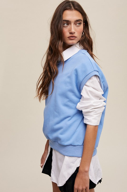 A woman stands against a neutral background, wearing the Short Sleeve Boxy Crew Neck Sweat Top in blue with ribbed detailing over a white button-up shirt.