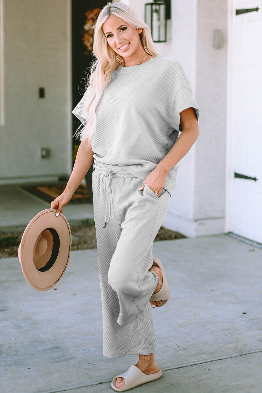 A woman with long hair smiles while standing outside a building, dressed in the Double Take Full Size Texture Short Sleeve Top and Pants Set, featuring a matching beige two-piece ensemble. She wears slides and holds a hat in her right hand. Her outfit, slightly stretchy for added comfort, is perfect for machine wash cold.