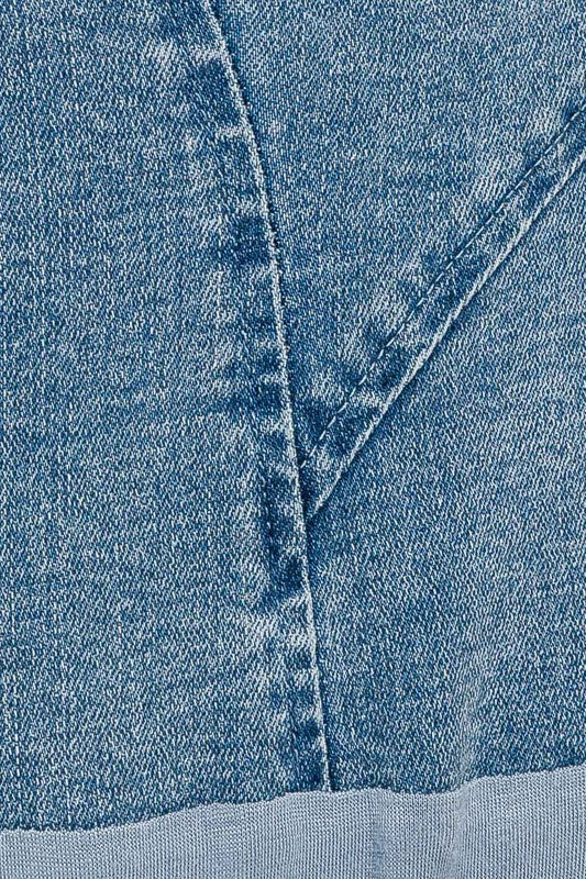 A woman with wavy brown hair models the Denim Bustier Bodysuit, featuring a sleek, fitted design and thin straps paired perfectly with a dark denim skirt adorned with pleats. She poses against a plain, light-colored background, drawing attention to her chic ensemble.