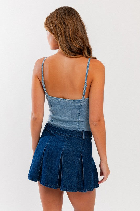 A woman with wavy brown hair models the Denim Bustier Bodysuit, featuring a sleek, fitted design and thin straps paired perfectly with a dark denim skirt adorned with pleats. She poses against a plain, light-colored background, drawing attention to her chic ensemble.