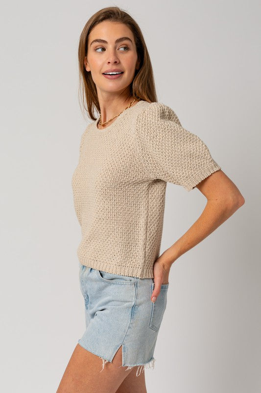 A woman in a straw hat and a Puff Sleeve Round Neck Texture Sweater Top holds a wicker bag with a pineapple. She's wearing light denim shorts, standing against a cream backdrop.