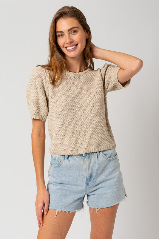 A woman in a straw hat and a Puff Sleeve Round Neck Texture Sweater Top holds a wicker bag with a pineapple. She's wearing light denim shorts, standing against a cream backdrop.