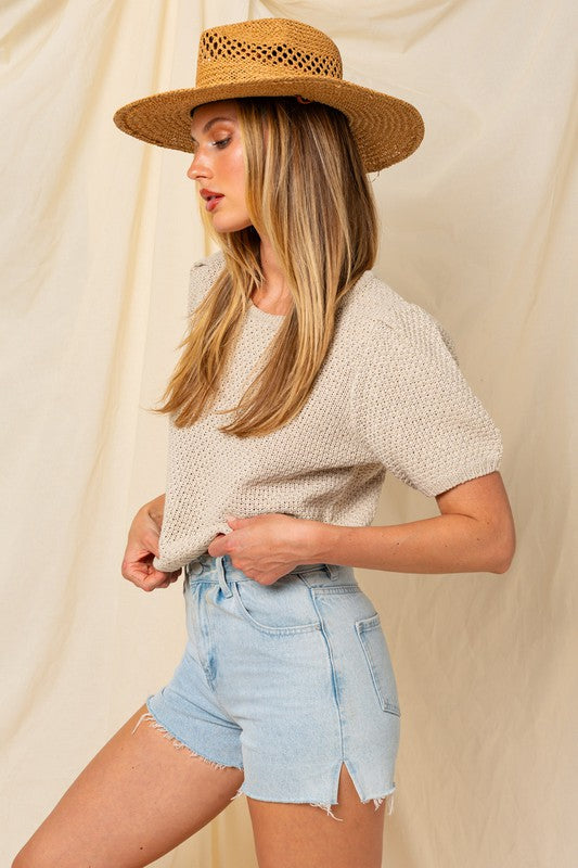 A woman in a straw hat and a Puff Sleeve Round Neck Texture Sweater Top holds a wicker bag with a pineapple. She's wearing light denim shorts, standing against a cream backdrop.