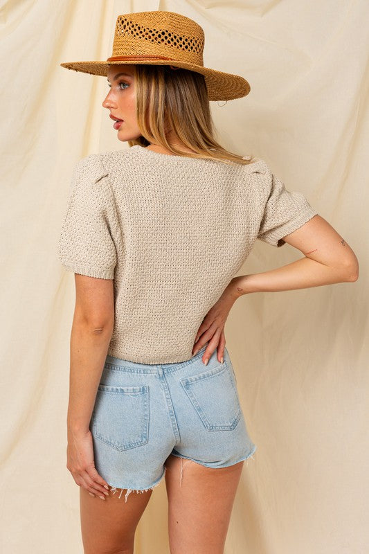 A woman in a straw hat and a Puff Sleeve Round Neck Texture Sweater Top holds a wicker bag with a pineapple. She's wearing light denim shorts, standing against a cream backdrop.