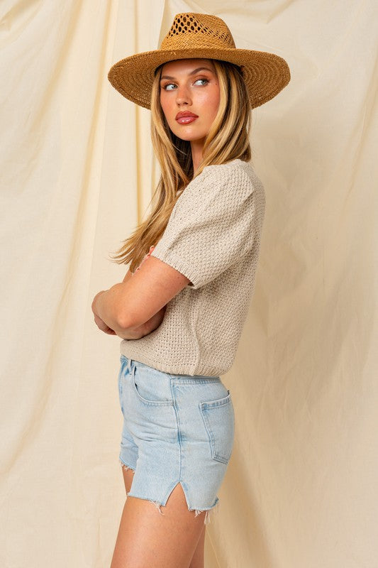 A woman in a straw hat and a Puff Sleeve Round Neck Texture Sweater Top holds a wicker bag with a pineapple. She's wearing light denim shorts, standing against a cream backdrop.