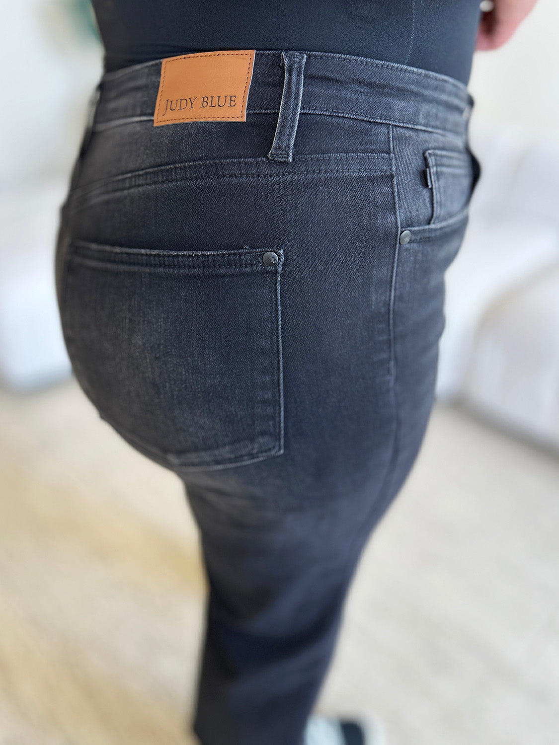 Person wearing Judy Blue Full Size High Waist Button Fly Jeans and black sneakers, standing indoors. Hands are in pockets, and the background includes a white couch and a plant. These high-waisted jeans make the outfit sleek and stylish.