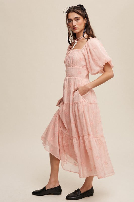 A woman strikes an elegant pose against a neutral background, showcasing the Flower Embroidered Puff Sleeve Tiered Maxi Dress by gracefully holding its tiered skirt.