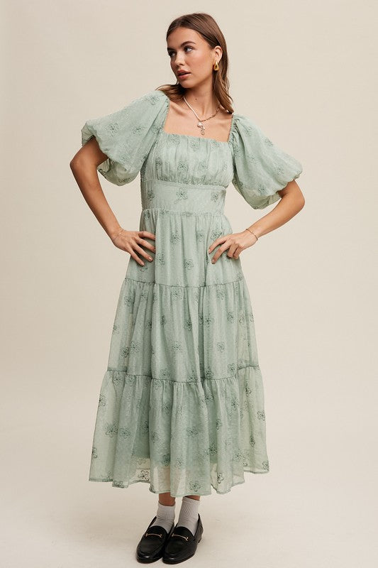 A woman strikes an elegant pose against a neutral background, showcasing the Flower Embroidered Puff Sleeve Tiered Maxi Dress by gracefully holding its tiered skirt.