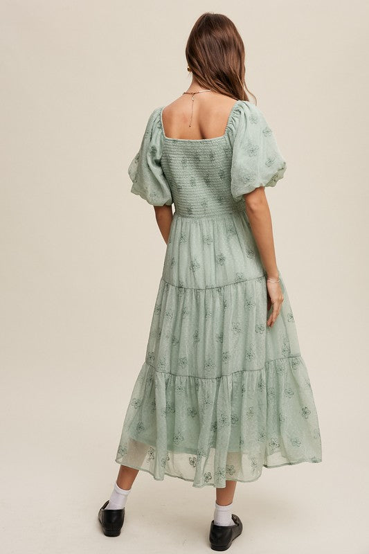 A woman strikes an elegant pose against a neutral background, showcasing the Flower Embroidered Puff Sleeve Tiered Maxi Dress by gracefully holding its tiered skirt.