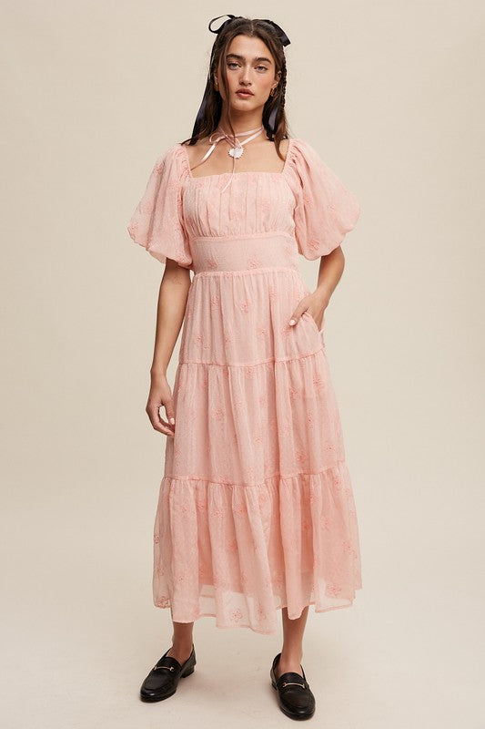 A woman strikes an elegant pose against a neutral background, showcasing the Flower Embroidered Puff Sleeve Tiered Maxi Dress by gracefully holding its tiered skirt.