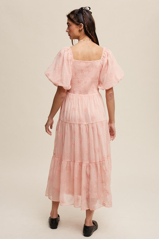 A woman strikes an elegant pose against a neutral background, showcasing the Flower Embroidered Puff Sleeve Tiered Maxi Dress by gracefully holding its tiered skirt.