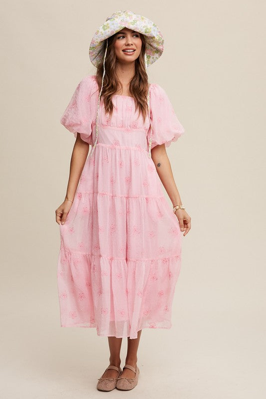 A woman strikes an elegant pose against a neutral background, showcasing the Flower Embroidered Puff Sleeve Tiered Maxi Dress by gracefully holding its tiered skirt.