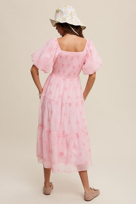 A woman strikes an elegant pose against a neutral background, showcasing the Flower Embroidered Puff Sleeve Tiered Maxi Dress by gracefully holding its tiered skirt.