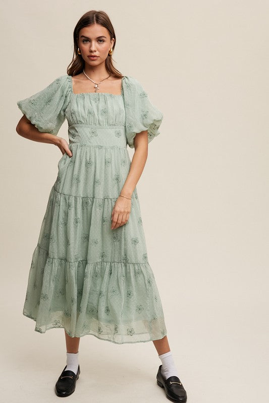 A woman strikes an elegant pose against a neutral background, showcasing the Flower Embroidered Puff Sleeve Tiered Maxi Dress by gracefully holding its tiered skirt.