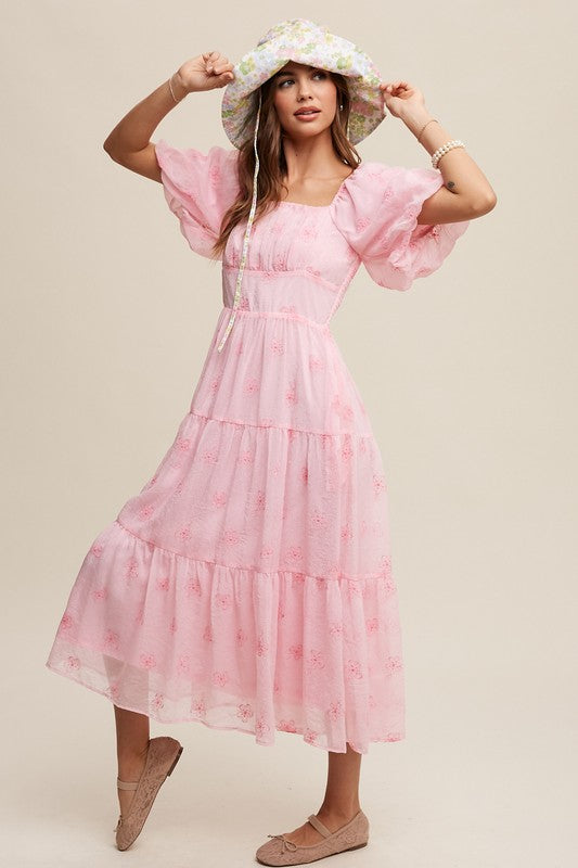 A woman strikes an elegant pose against a neutral background, showcasing the Flower Embroidered Puff Sleeve Tiered Maxi Dress by gracefully holding its tiered skirt.