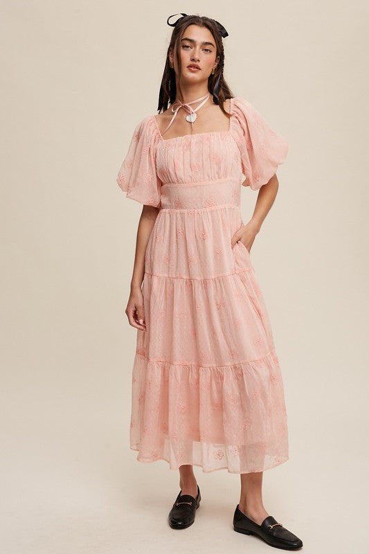 A woman strikes an elegant pose against a neutral background, showcasing the Flower Embroidered Puff Sleeve Tiered Maxi Dress by gracefully holding its tiered skirt.