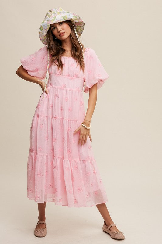 A woman strikes an elegant pose against a neutral background, showcasing the Flower Embroidered Puff Sleeve Tiered Maxi Dress by gracefully holding its tiered skirt.