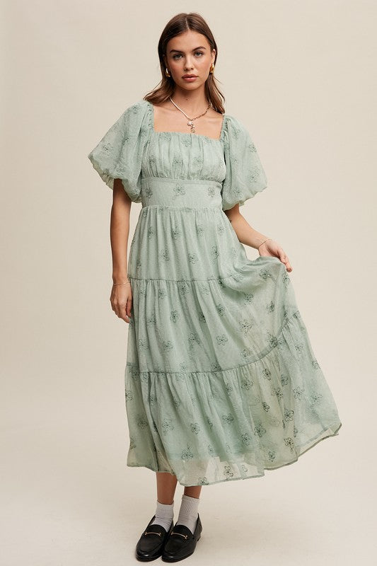 A woman strikes an elegant pose against a neutral background, showcasing the Flower Embroidered Puff Sleeve Tiered Maxi Dress by gracefully holding its tiered skirt.