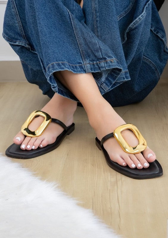 Close-up of feet in AVA 4 smooth leather tan sandals with large gold buckles, standing on a plush, light gray carpet. The person is wearing denim jeans.