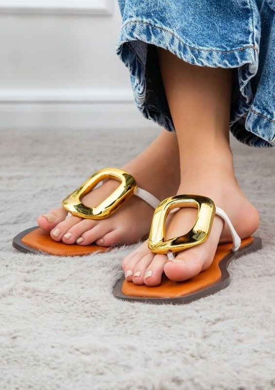 Close-up of feet in AVA 4 smooth leather tan sandals with large gold buckles, standing on a plush, light gray carpet. The person is wearing denim jeans.