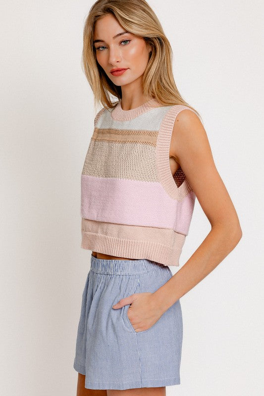 A woman wearing a Round Neck Sweater Vest with pastel stripes stands against a plain background. She has long, light brown hair and pairs her outfit with light blue striped pants, embracing the sleeveless style in a chic yet casual manner.