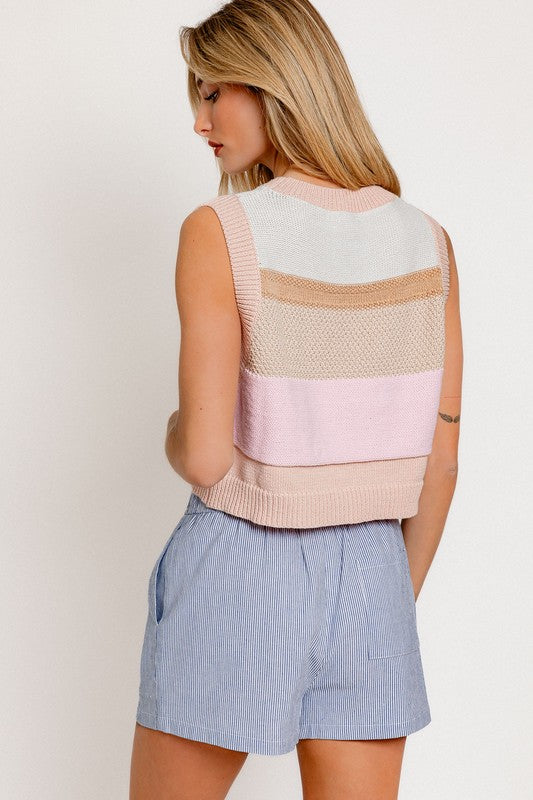 A woman wearing a Round Neck Sweater Vest with pastel stripes stands against a plain background. She has long, light brown hair and pairs her outfit with light blue striped pants, embracing the sleeveless style in a chic yet casual manner.