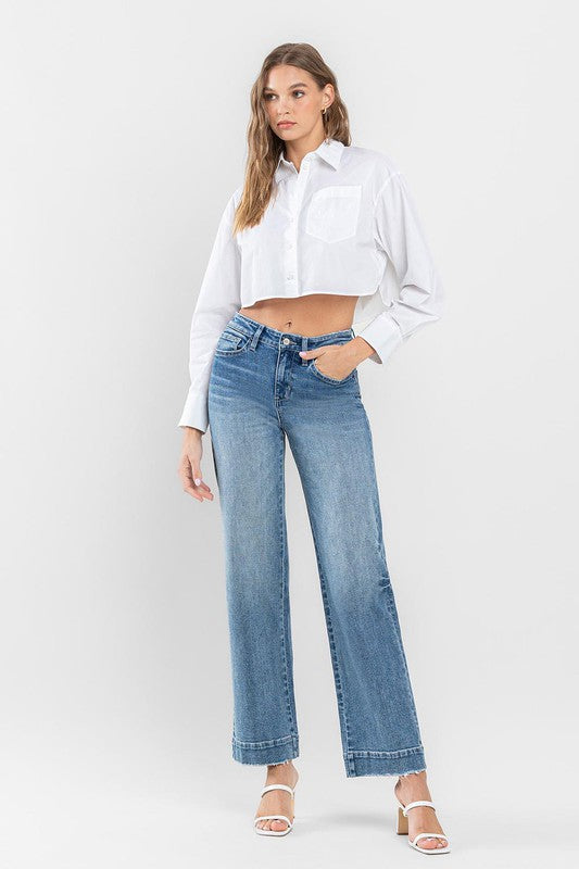 A woman in a white tank top and High Rise Wide Leg Jeans with Trouser Hem Detail sits casually on an armchair with one leg draped over the arm, indoors near a glass door with a view of greenery outside.