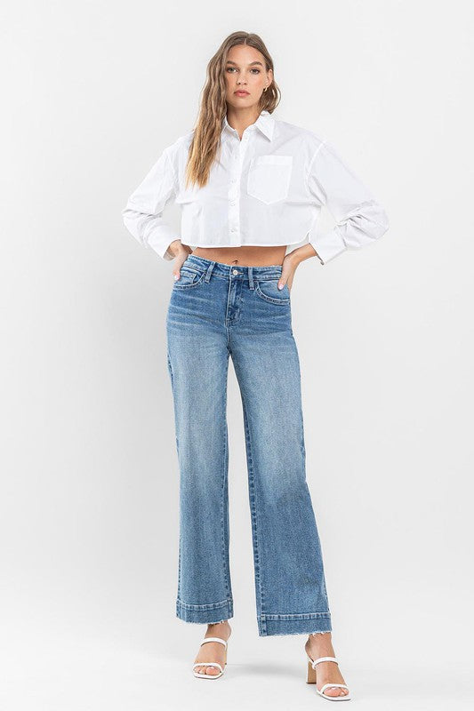 A woman in a white tank top and High Rise Wide Leg Jeans with Trouser Hem Detail sits casually on an armchair with one leg draped over the arm, indoors near a glass door with a view of greenery outside.