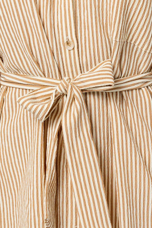 A woman wearing the Half Sleeve Button Down Shirt Dress, featuring a beige and white striped pattern, exudes casual elegance as she stands confidently against a plain background. The design is enhanced by half-sleeves and a tied waist belt, which perfectly complement her style.