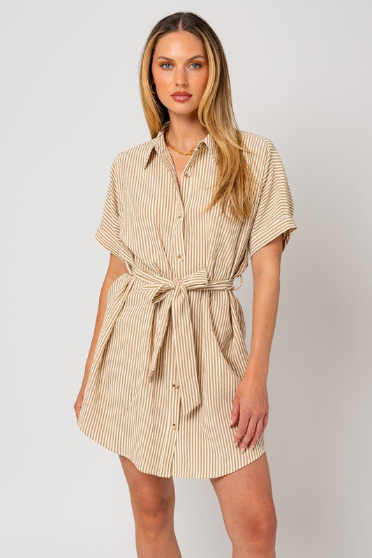 A woman wearing the Half Sleeve Button Down Shirt Dress, featuring a beige and white striped pattern, exudes casual elegance as she stands confidently against a plain background. The design is enhanced by half-sleeves and a tied waist belt, which perfectly complement her style.