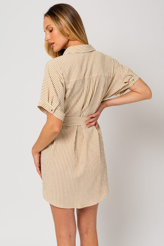 A woman wearing the Half Sleeve Button Down Shirt Dress, featuring a beige and white striped pattern, exudes casual elegance as she stands confidently against a plain background. The design is enhanced by half-sleeves and a tied waist belt, which perfectly complement her style.