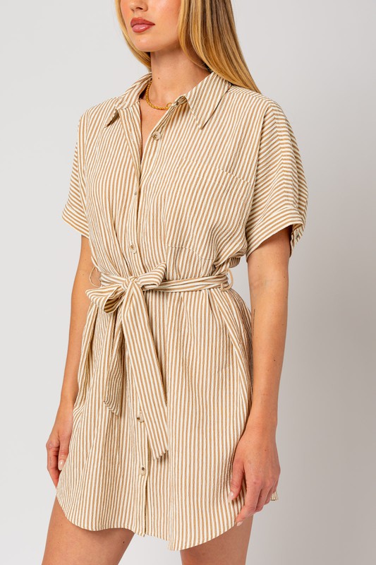 A woman wearing the Half Sleeve Button Down Shirt Dress, featuring a beige and white striped pattern, exudes casual elegance as she stands confidently against a plain background. The design is enhanced by half-sleeves and a tied waist belt, which perfectly complement her style.