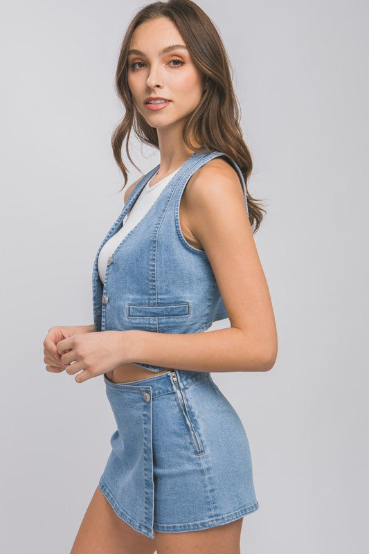 A woman wearing a Denim Buttoned Vest Top over a white shirt and a denim mini skirt stands against a plain background with one hand on her hip and the other by her side.