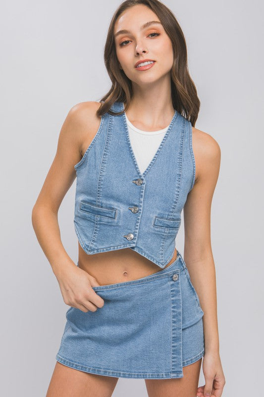 A woman wearing a Denim Buttoned Vest Top over a white shirt and a denim mini skirt stands against a plain background with one hand on her hip and the other by her side.