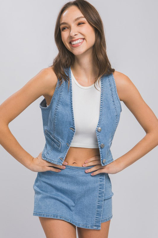 A woman wearing a Denim Buttoned Vest Top over a white shirt and a denim mini skirt stands against a plain background with one hand on her hip and the other by her side.