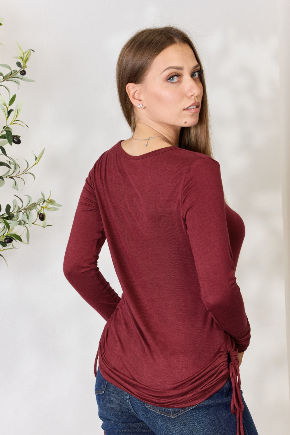 A woman stands in front of a plain background and a small tree, wearing the Culture Code Full Size Drawstring Round Neck Long Sleeve Top in maroon and blue jeans.